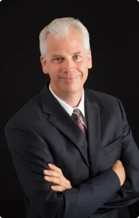 Professional headshot of a smiling person in a dark suit with arms crossed on a black background