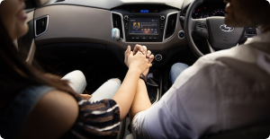 Two people holding hands in a car, viewed from the back seat with the dashboard ahead.
