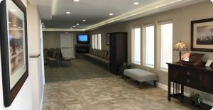 Modern hallway lounge with carpet, bench, framed art, desk, and chairs near a TV at the far end