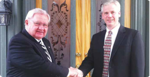 Two men in suits shaking hands indoors in front of ornate doors and gold curtains.