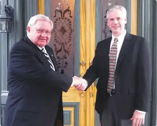 Two men in suits shaking hands in front of an ornate yellow doorway.