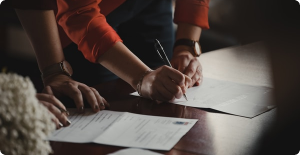 Hands signing a document at a wooden table with papers and a pen in warm indoor light