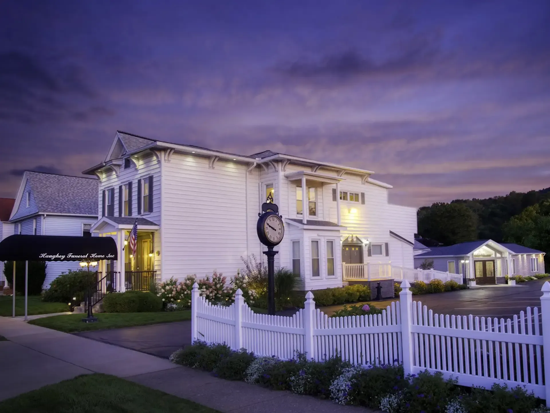 White Victorian-style building with lit windows and a white picket fence at dusk