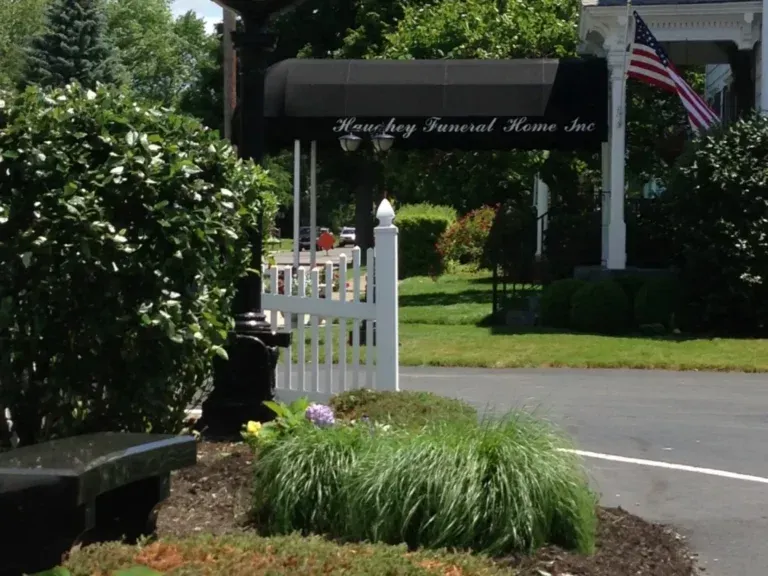 Garden entrance with white gate, black awning, U.S. flag, and lush greenery beside a driveway.
