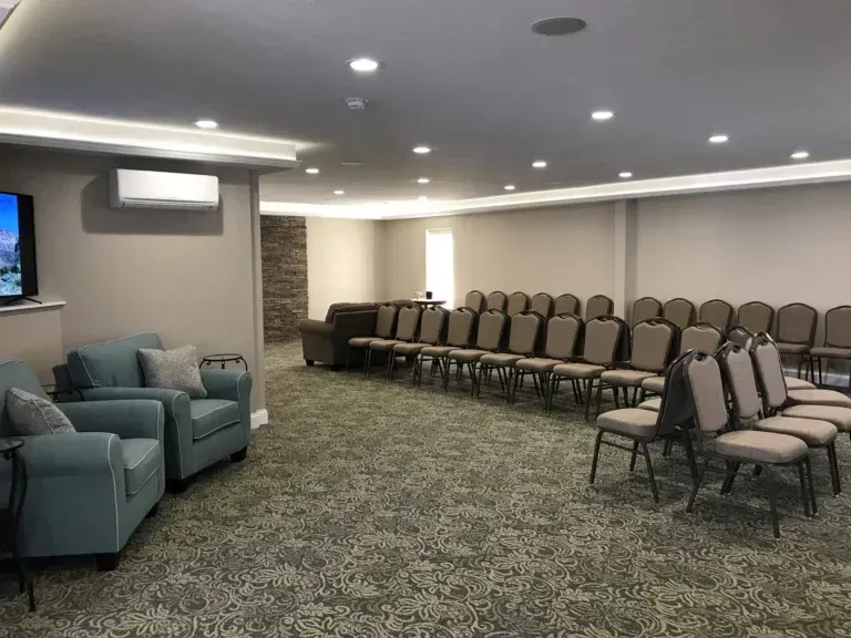 Empty waiting room with teal chairs, rows of tan folding chairs, and patterned carpet under recessed lights