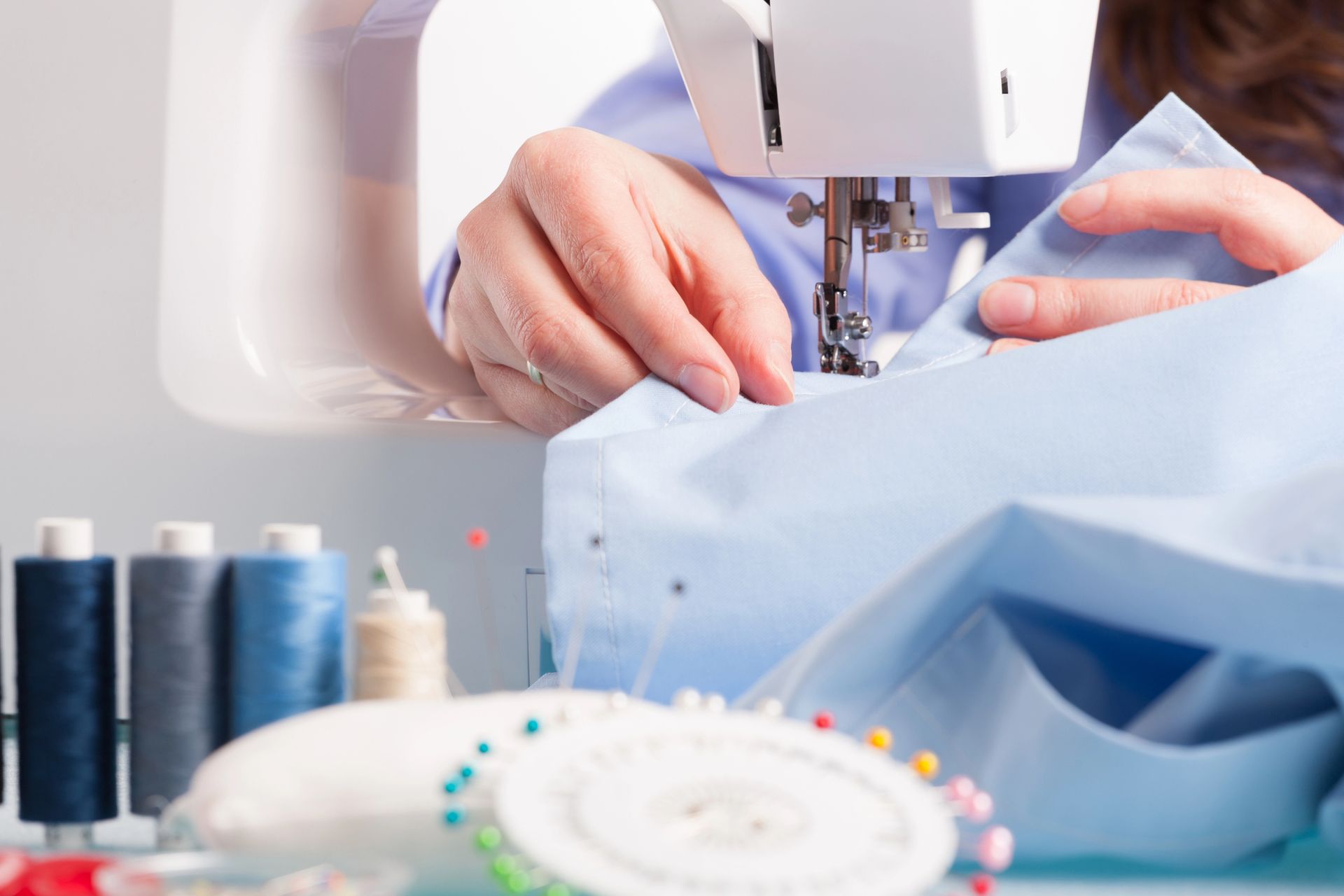 Una persona cose una camisa azul claro con una máquina de coser blanca. En primer plano se ven carretes de hilo y alfileres.