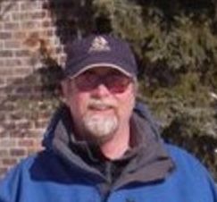 A man wearing a blue jacket and a hat is standing in front of a brick wall.