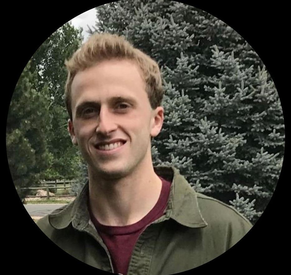 A man in a green jacket is smiling in front of a pine tree