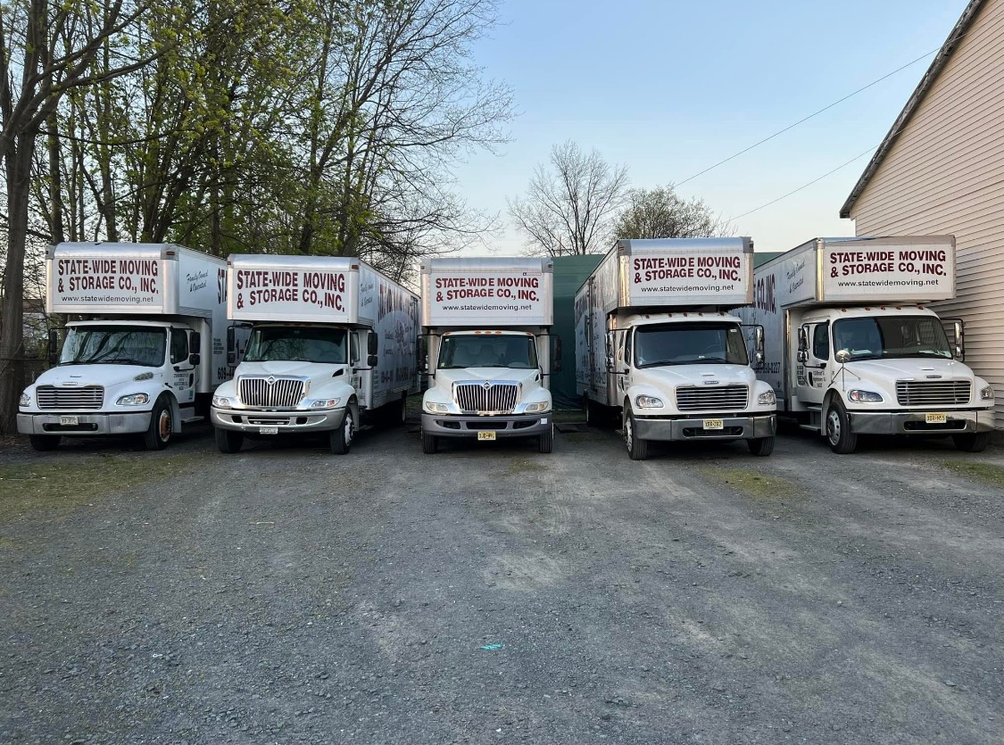 Five white box trucks parked in front of a building on gravel. The trucks have the same logo.