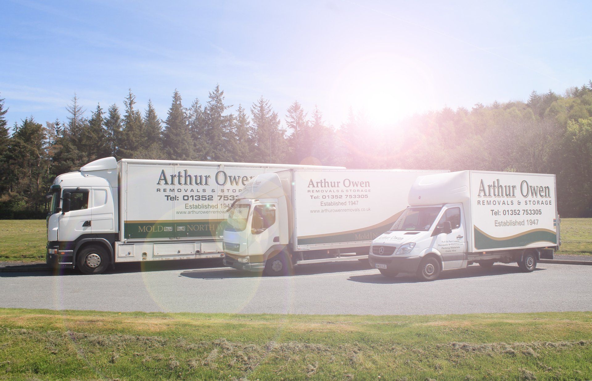 Two Arthur Owen Removals & Storage moving trucks parked outdoors on a sunny roadside with trees in the background, alongside a Arthur Owen Removals van.