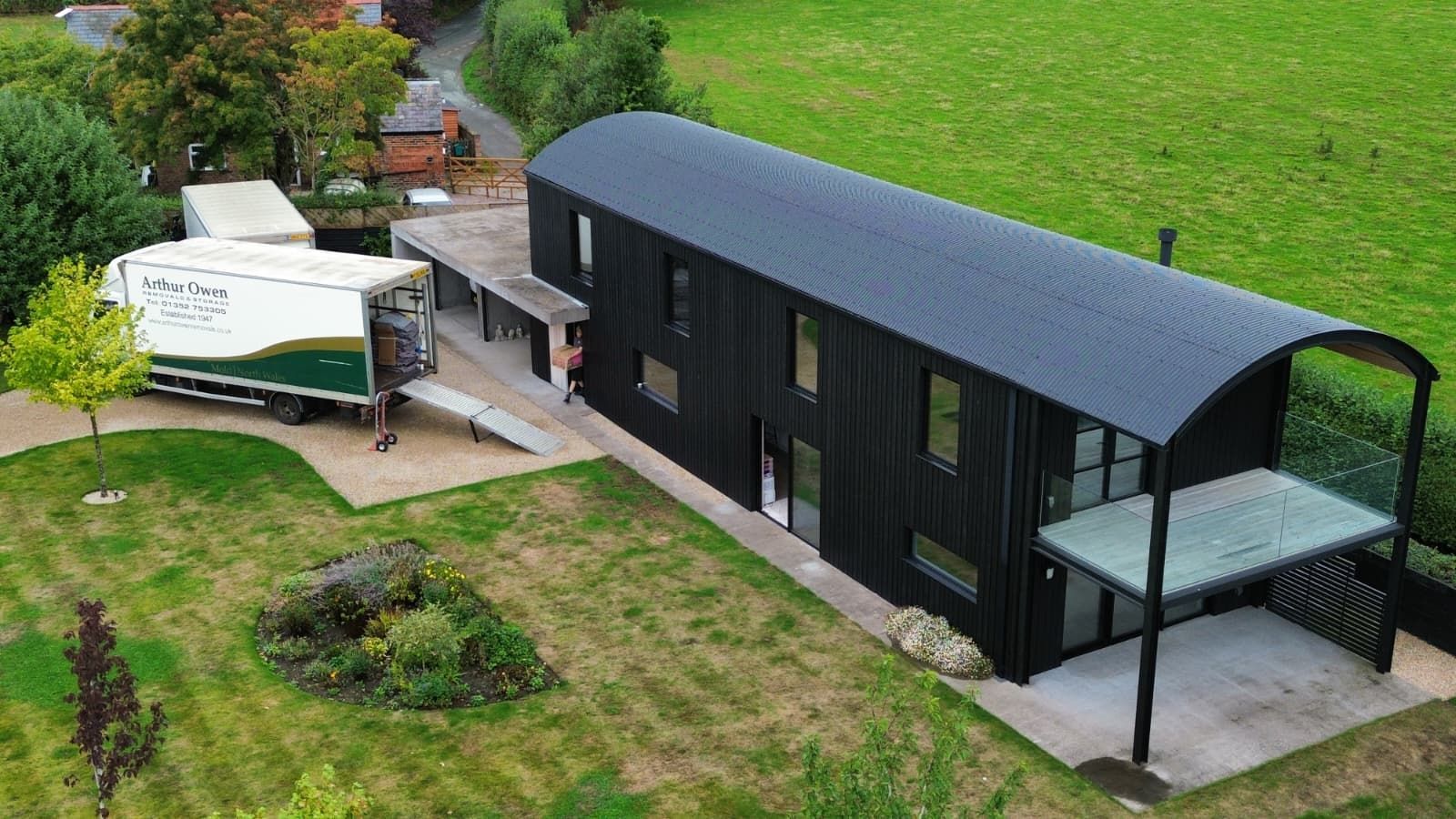 Black modern architectural house on a grassy lot with a white Arthur Owen Removals truck alongside loading, seen from above.