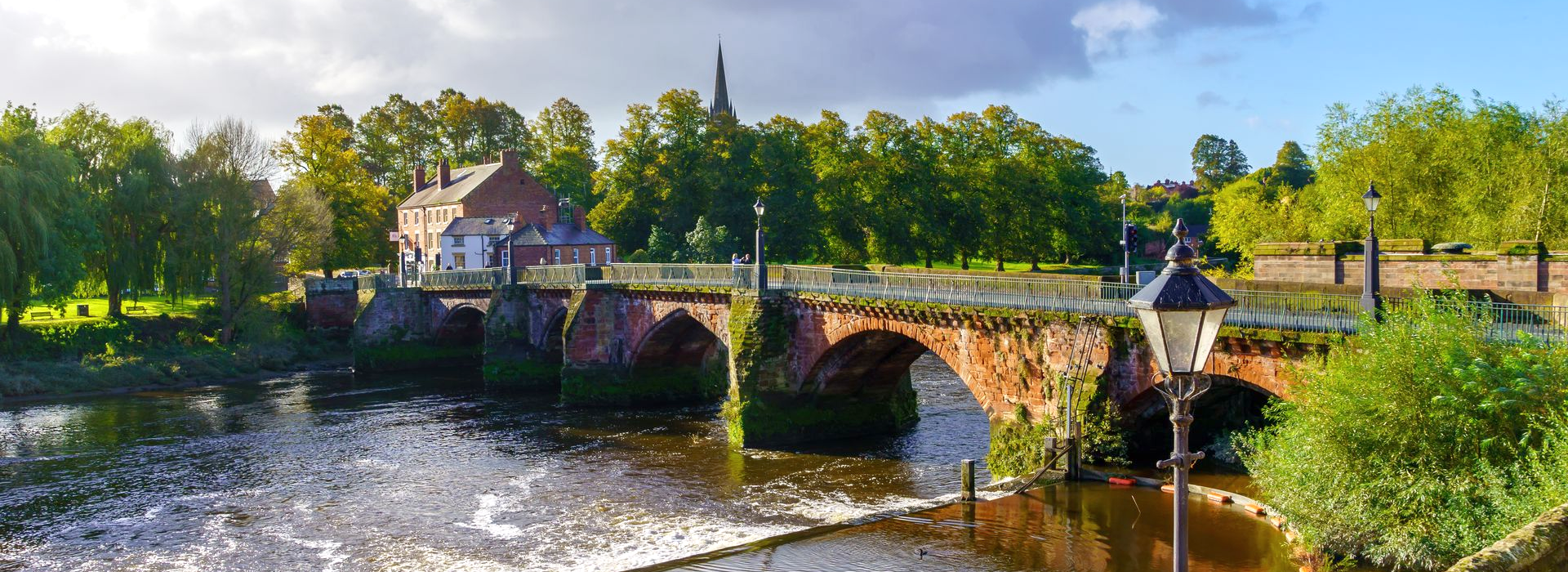 Stone bridge over the River Dee, Chester beside a waterfall, with trees and a partly cloudy sky.
