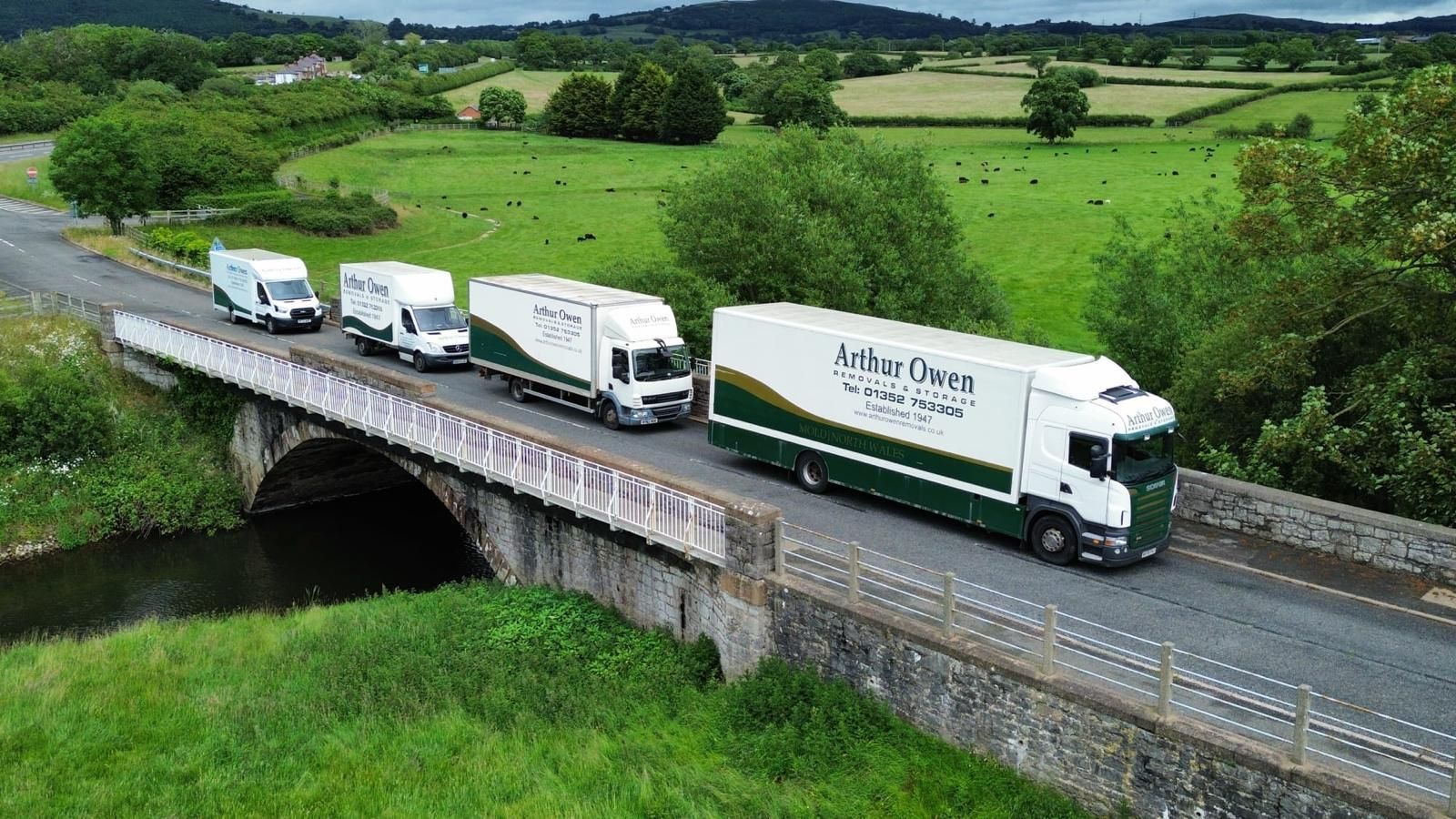 Two Arthur Owen Removals & Storage moving trucks parked outdoors on a sunny roadside with trees in the background, alongside a Arthur Owen Removals van.