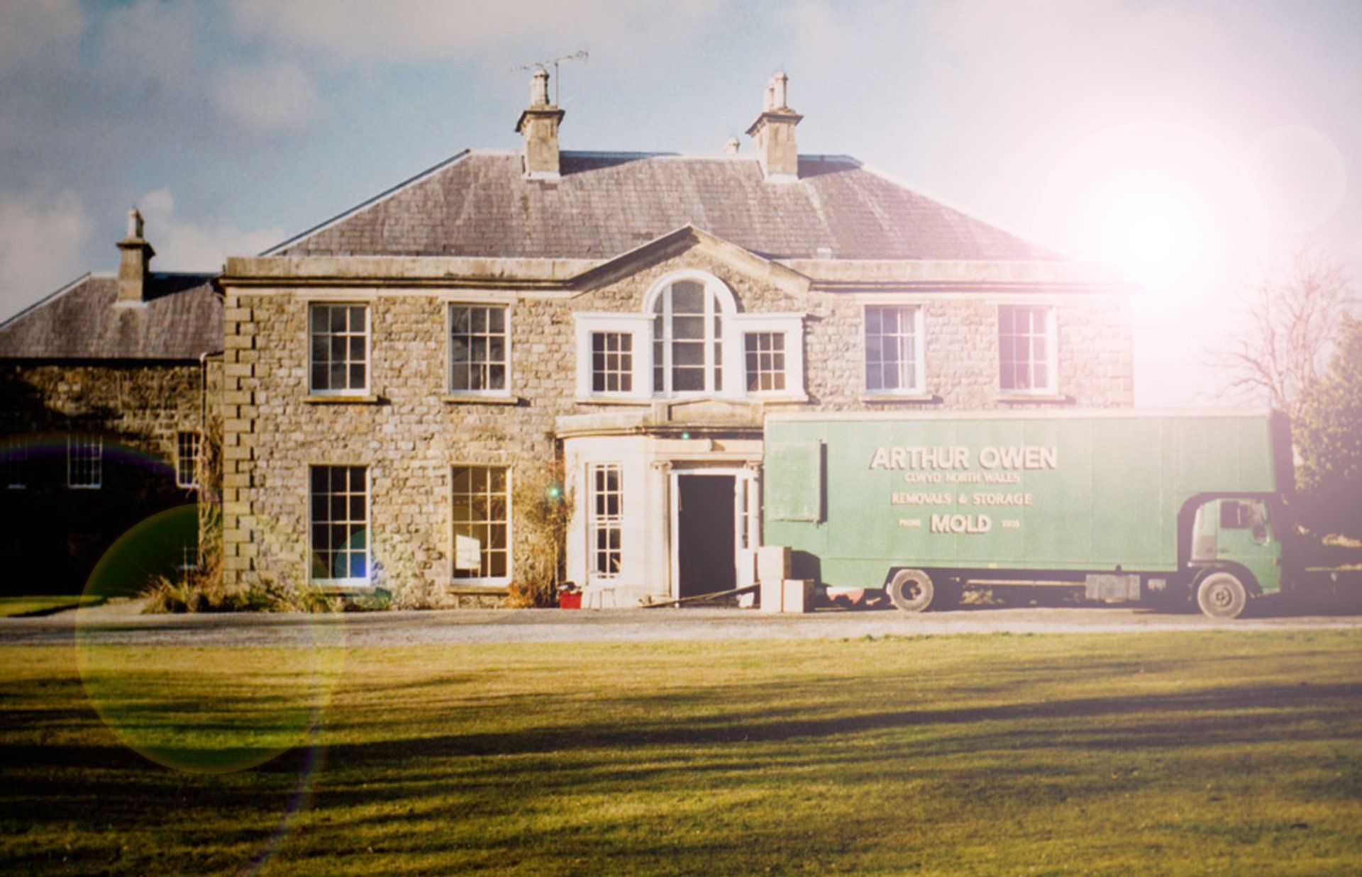 Stone house in sunlight with a green Arthur Owen moving truck parked outside on a grassy lawn.