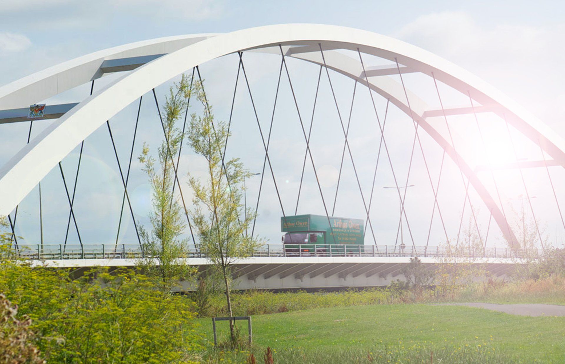 White arched bridge over a grassy field with trees and bright sun glare and an Arthur Owen Removals truck driving over the bridge.