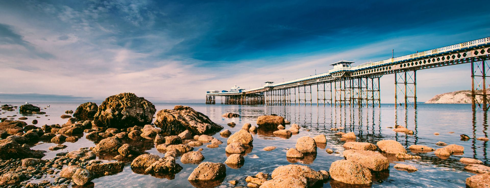 Rocky shoreline at sunset with a long view of Llandudno Pier stretching across calm water and a blue sky