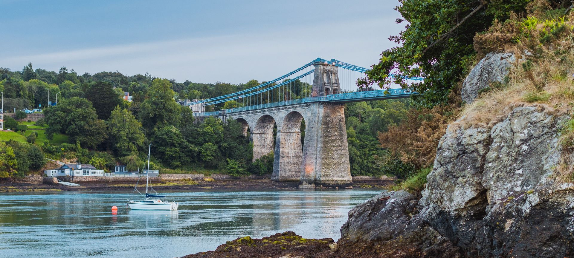 Menai suspension bridge over the Menai Straits, Anglesey beside rocky cliffs and green trees, with a sailboat in the water.