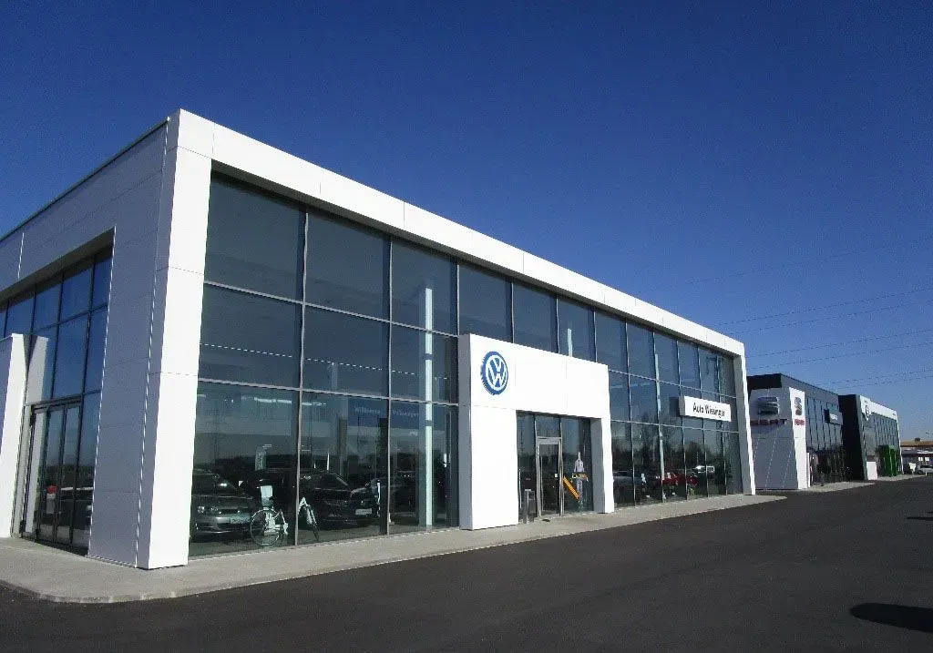 Modernes Autohausgebäude mit Glasfassade und blauem Logo, vom Parkplatz aus gesehen.