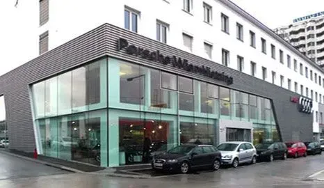 Modernes Gebäude mit Glasfassade an einer regnerischen Straße mit geparkten Autos und einer weißen oberen Fassade