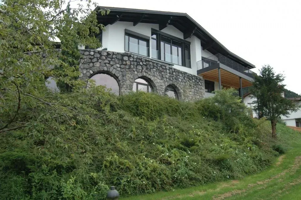 Steinhaus an einem grünen Hang mit Bogenfenstern und schrägem Dach, umgeben von dichter Vegetation.