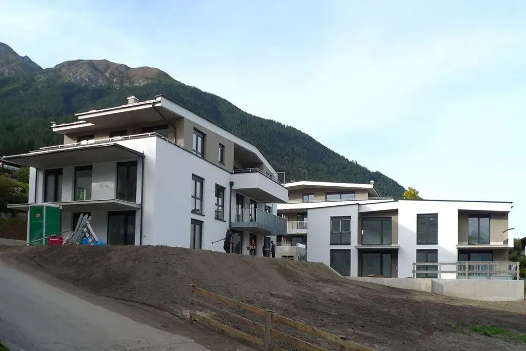 Moderne, weiße Hanghäuser mit Balkonen neben einer Schotterstraße, im Hintergrund Berge