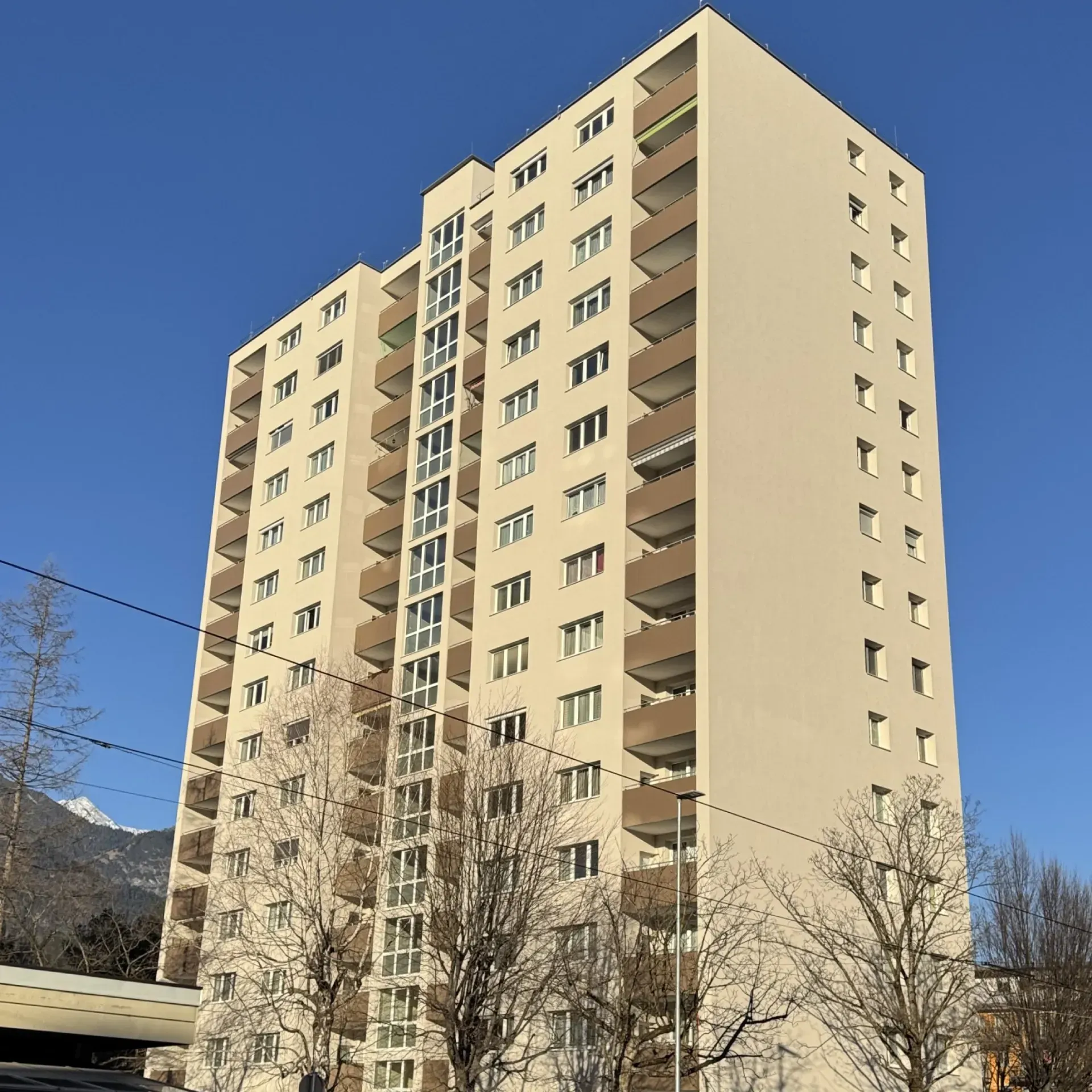 Beigefarbenes Hochhaus vor klarem, blauem Himmel, davor kahle Bäume.