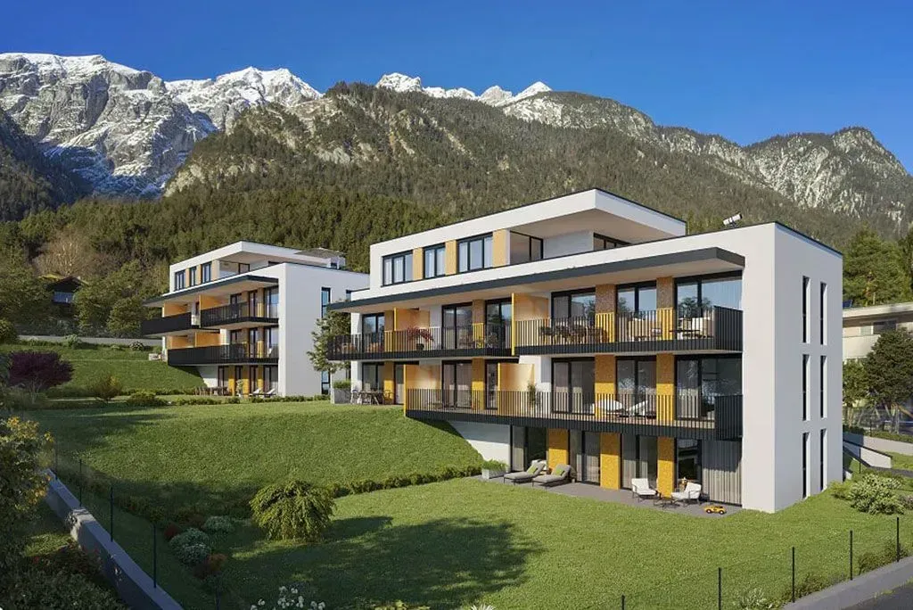 Moderne Apartmentgebäude mit Balkonen in grüner Berglandschaft, im Hintergrund schneebedeckte Gipfel