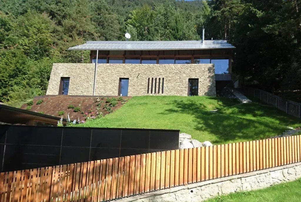 Steinhaus an einem grünen Hang mit Holzzaun und Bäumen im Hintergrund