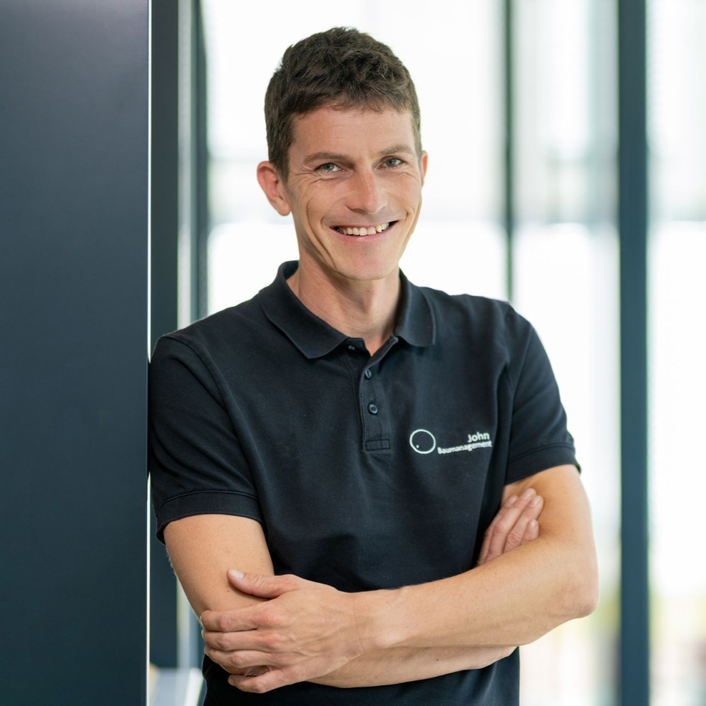 Ein lächelnder Mann in einem schwarzen Poloshirt steht mit verschränkten Armen in einem hellen Büro.