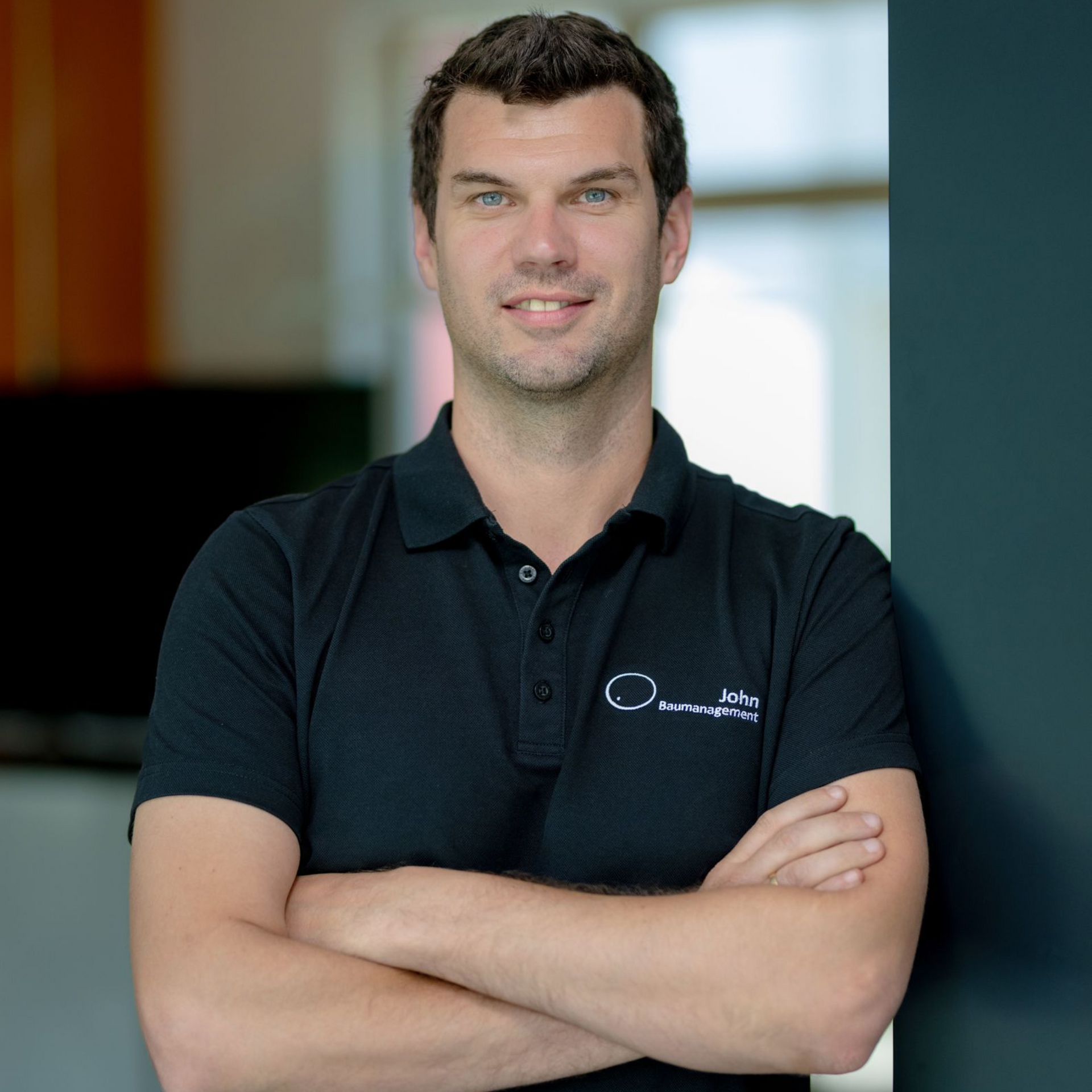 Ein Mann mit verschränkten Armen steht in einem verschwommenen Innenraum und trägt ein schwarzes Poloshirt.