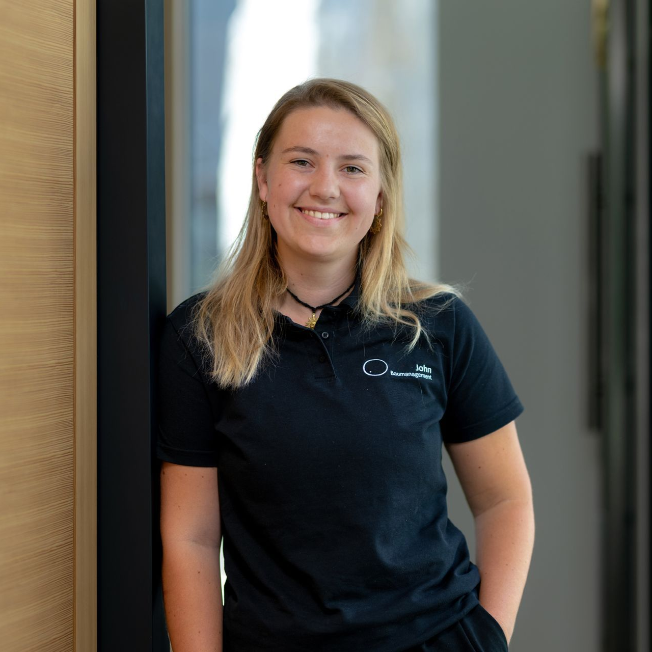 Eine lächelnde Frau in einem schwarzen Poloshirt steht drinnen in einem Flur.