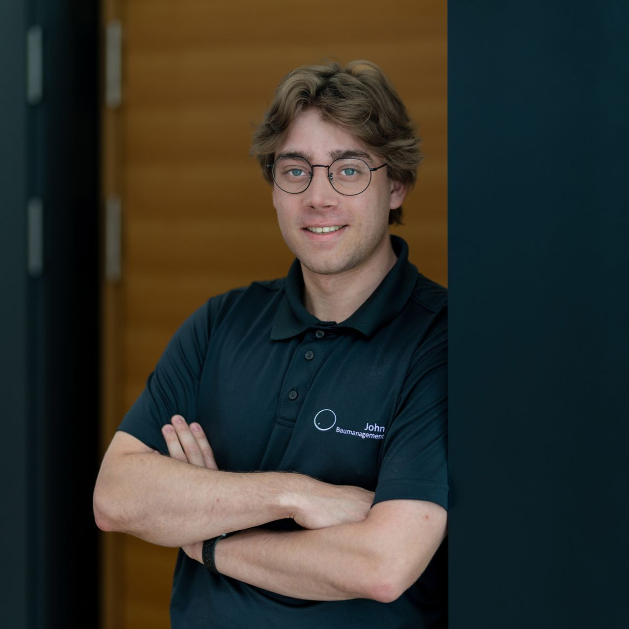 Eine lächelnde Person mit Brille und verschränkten Armen in einem dunklen Poloshirt steht in einem Türrahmen.