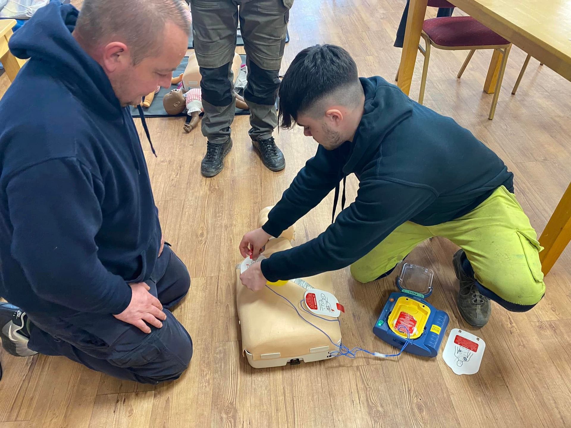 Two men practice using an AED on a mannequin in a room. One man kneels, watching; the other applies pads to the dummy.