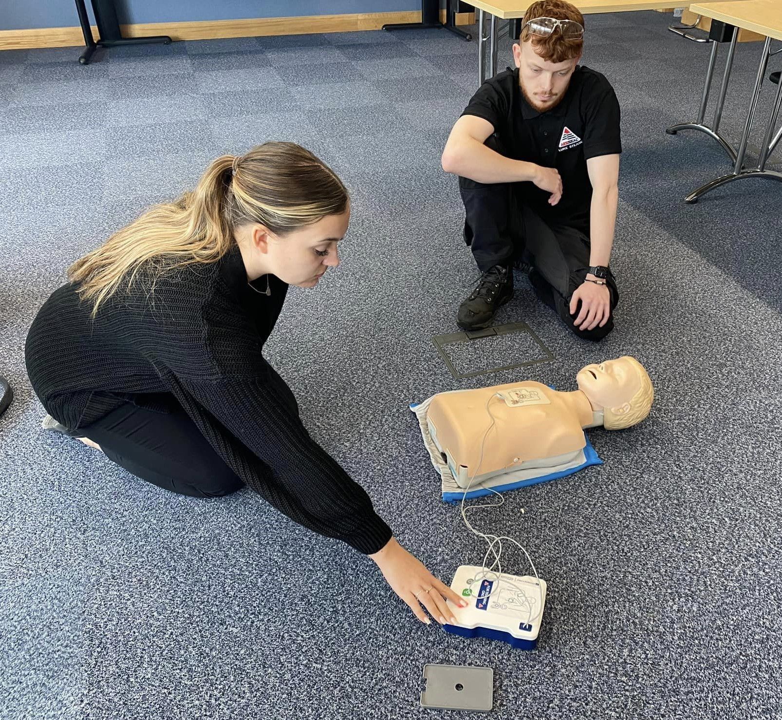 Two people practice CPR on a training dummy, one kneels operating an AED. They are on a carpeted floor in a room.