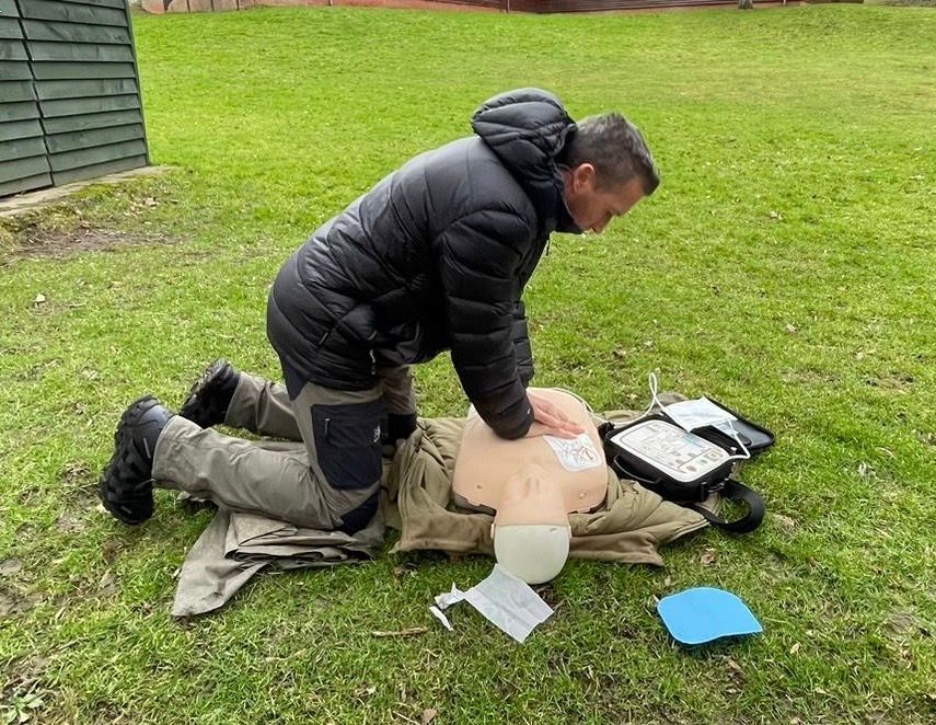Person performing CPR on a training dummy outdoors on green grass. The person is kneeling, pressing on the dummy's chest.