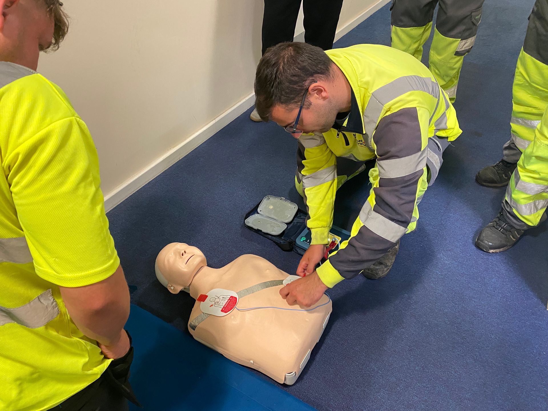 Mannequin receiving AED training. A man kneels and applies pads to a training mannequin. Other trainees watch.