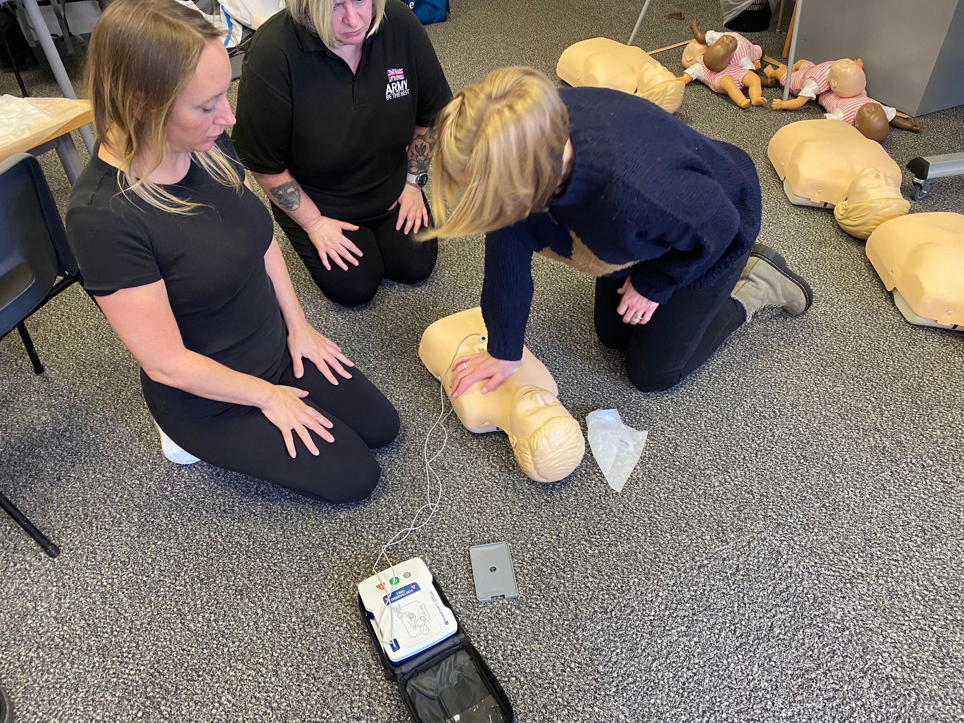 Three people practicing CPR on a training dummy. One person is kneeling, performing chest compressions while the other two watch. An AED is on the floor.