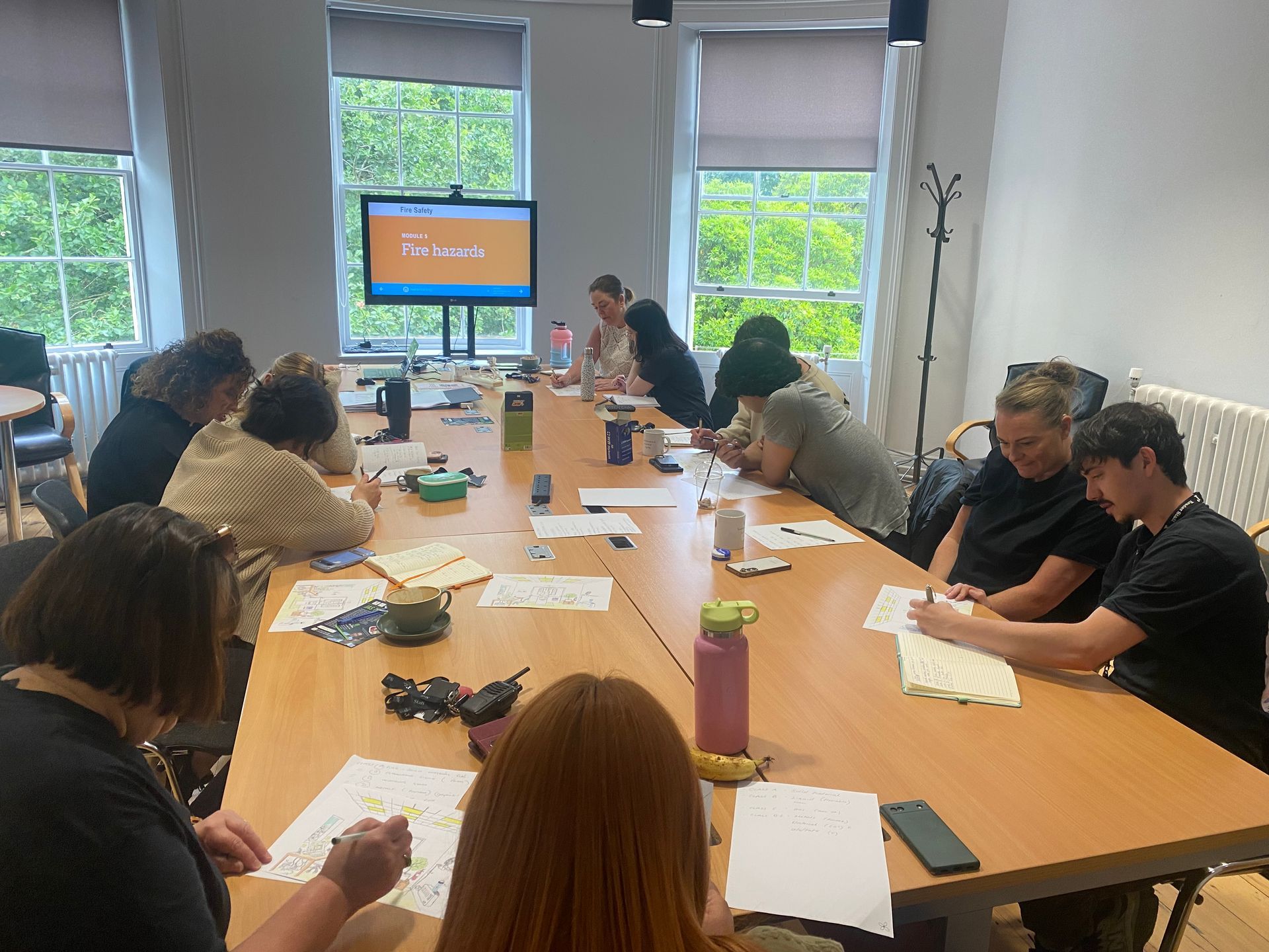 People at a long table in a well-lit room are drawing. A screen displays the word