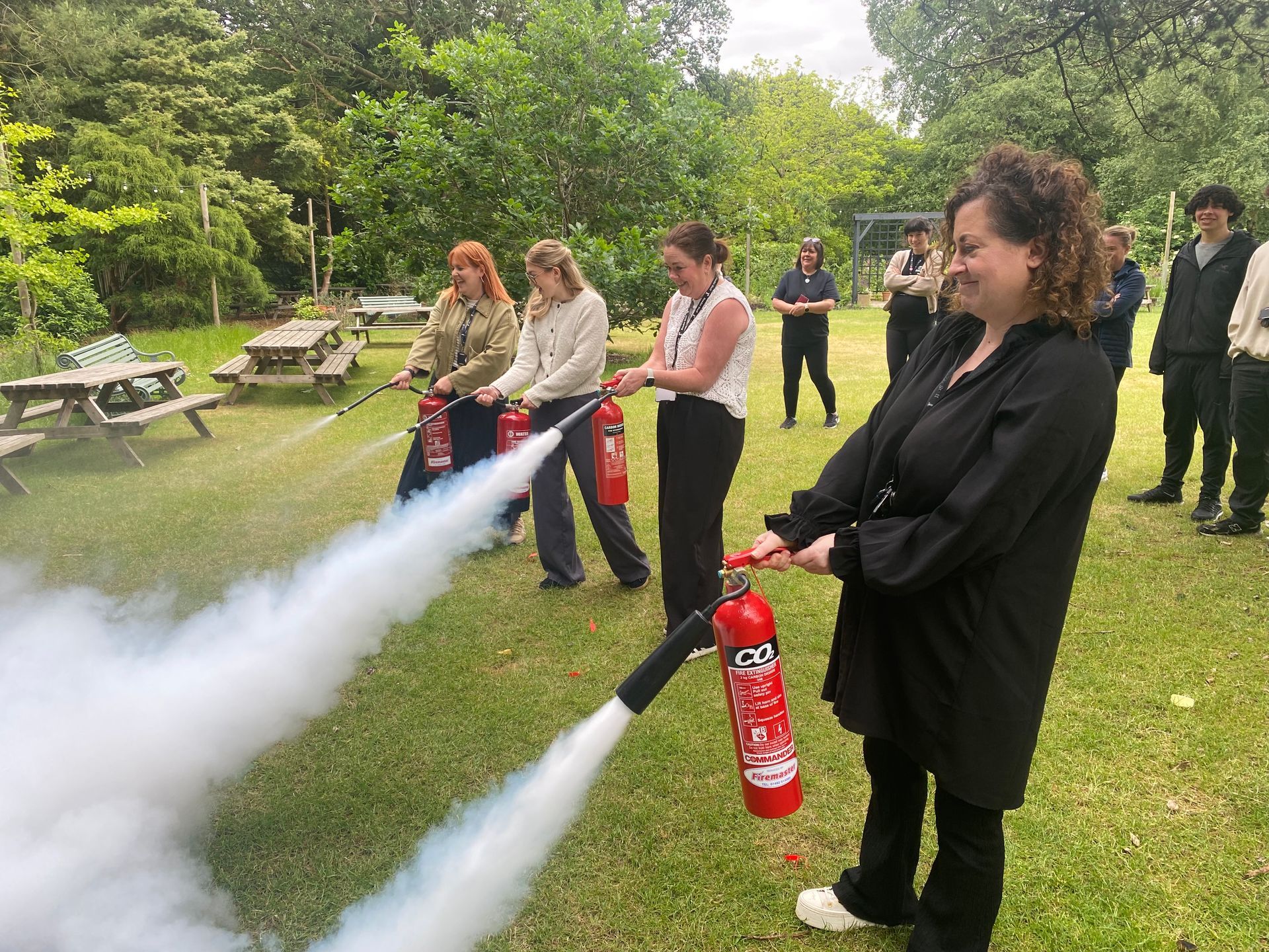 Group of people practicing fire extinguisher use outdoors. Several people spray extinguishers, creating white plumes of smoke.