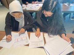 Two women collaborating, pointing at documents on a table. One wears a white hijab. They are in a well-lit room.
