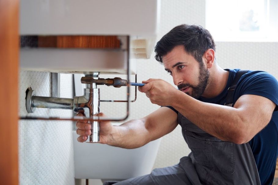 Plumber repairing a pipe under a kitchen sink with a wrench, crouched beside the cabinet.