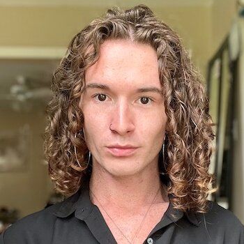 A young man with long curly hair is wearing a black shirt and earrings.