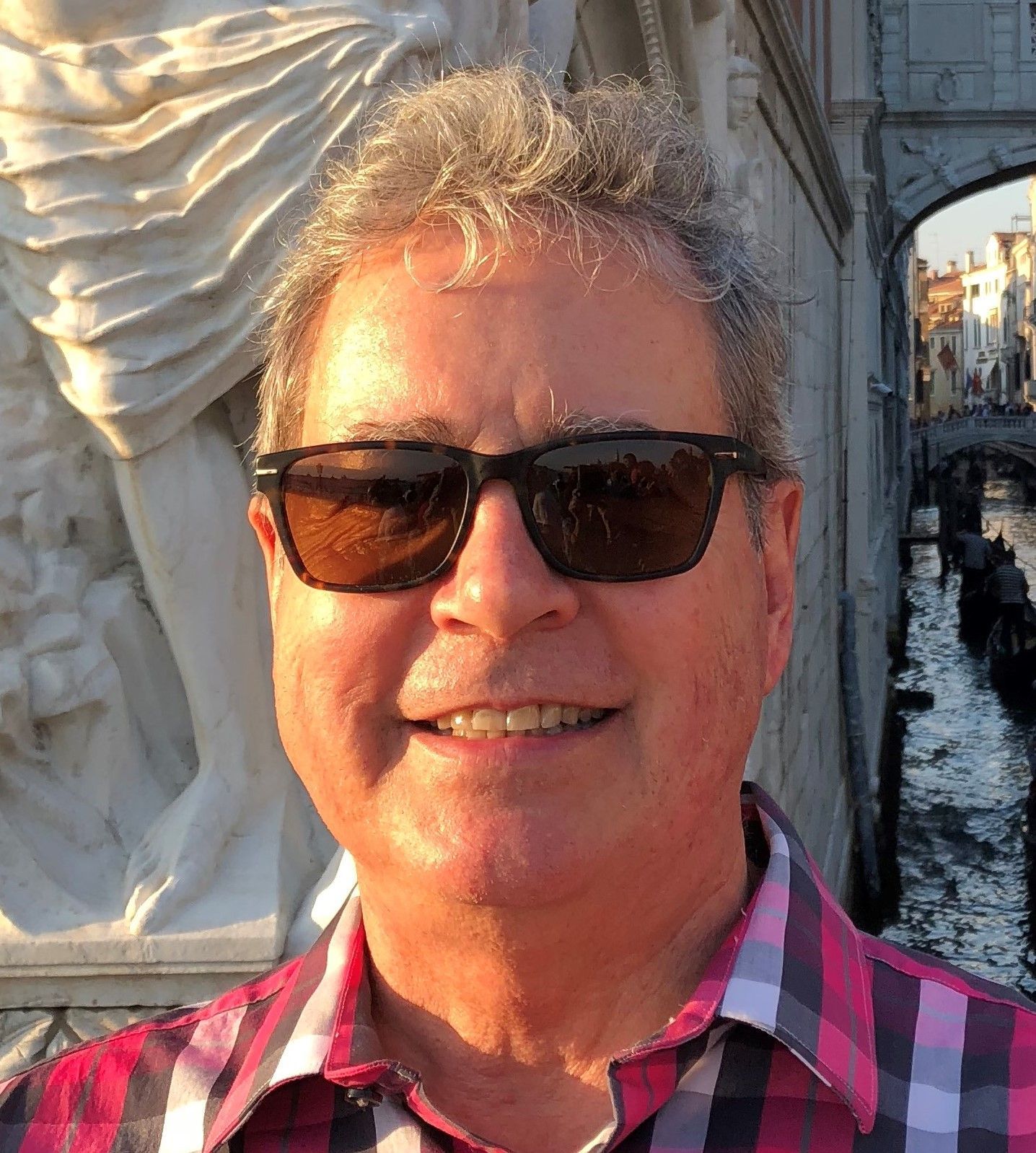 A man wearing sunglasses and a plaid shirt is smiling in front of a statue.