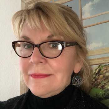 A woman wearing glasses and earrings is standing in front of a painting.
