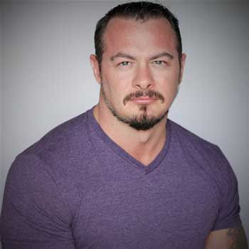 A man with a beard is wearing a purple shirt and looking at the camera.