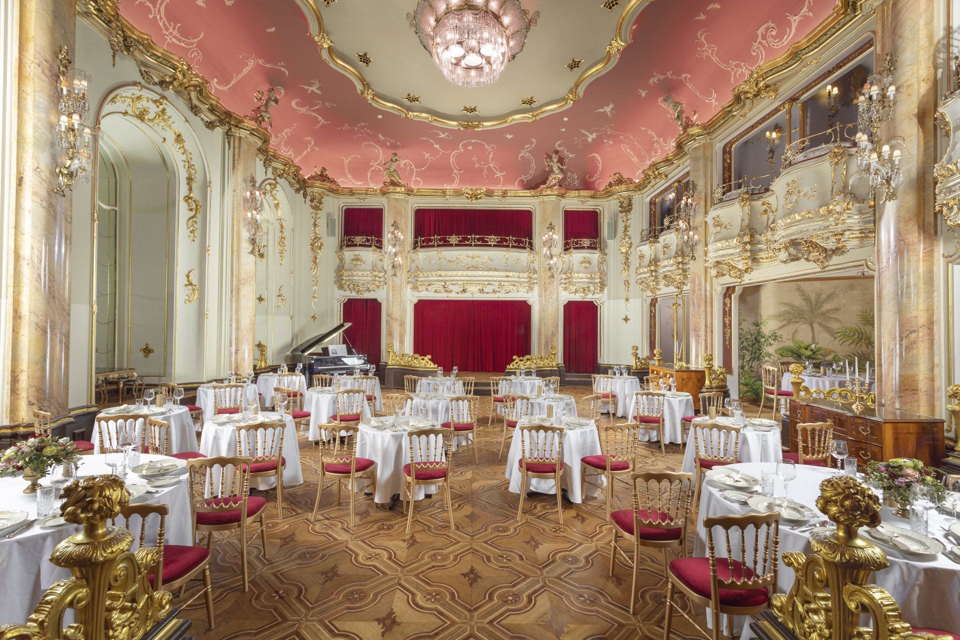 Elegant banquet hall with gold décor, pink ceiling, and round tables set with white linens and red chairs