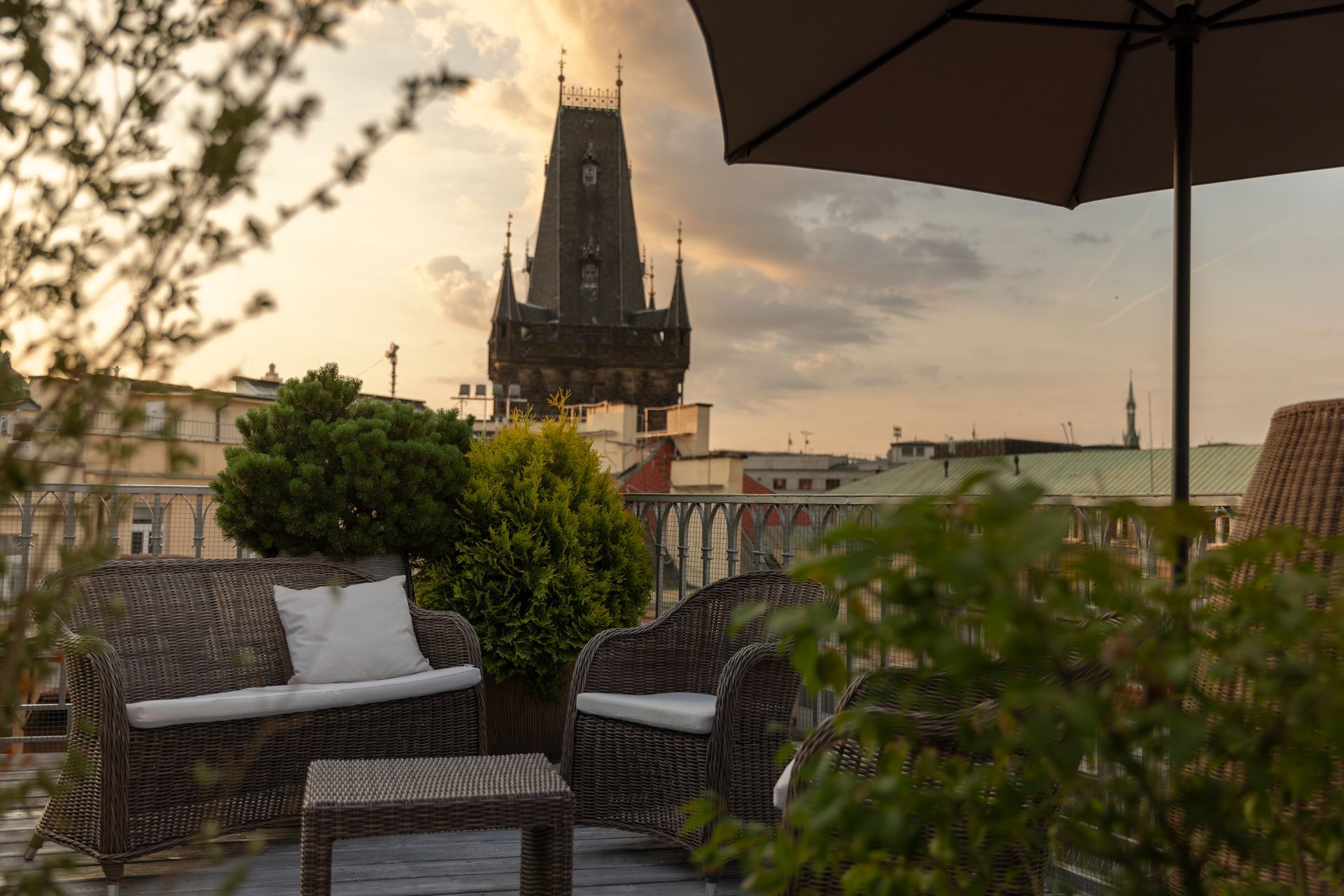 Střešní terasa s proutěnými židlemi a panoramatem města při západu slunce, v dálce vysoká věž katedrály