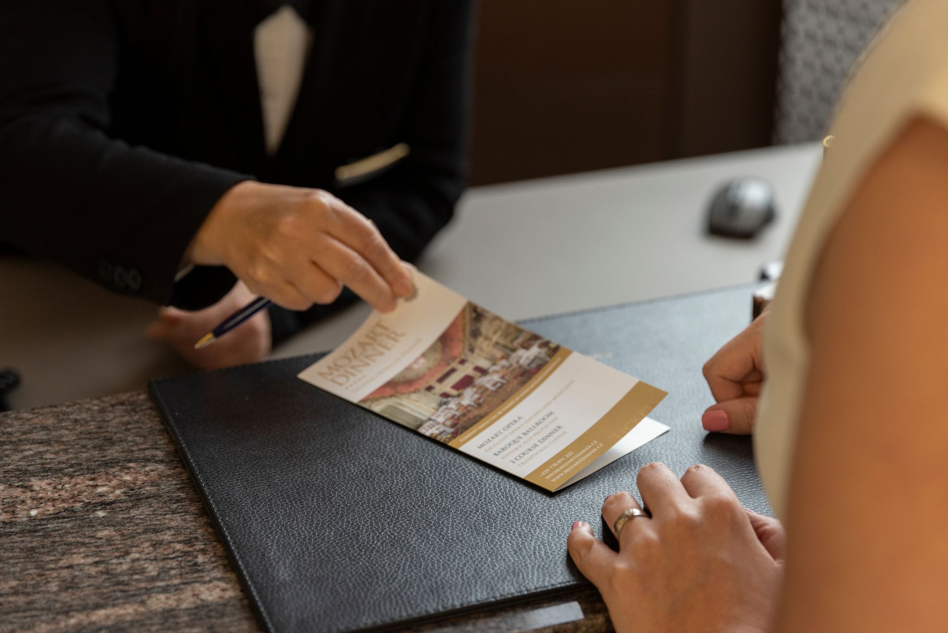 Hands exchanging a brochure across a desk beside a black portfolio folder.