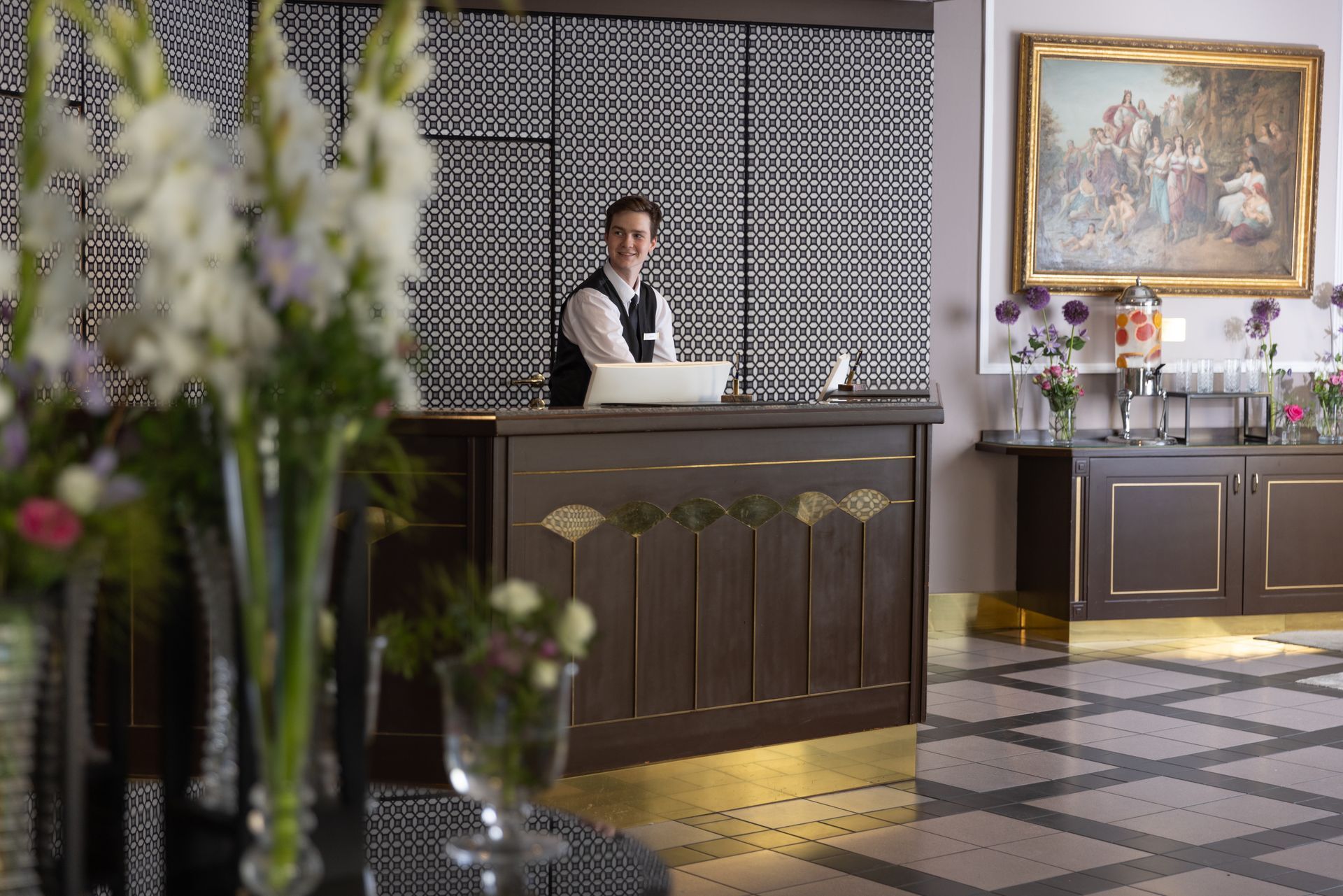 Hotel reception desk with a clerk, floral arrangements, and a large framed painting on the wall
