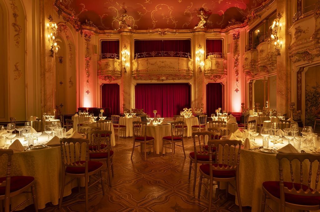 Elegant banquet hall with candlelit tables, ornate gold decor, and a red stage curtain